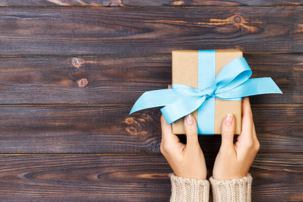 Woman hands give wrapped valentine or other holiday handmade present in paper with red, blue and green ribbon. Present box, decoration of gift on white wooden table, top view with copy space.