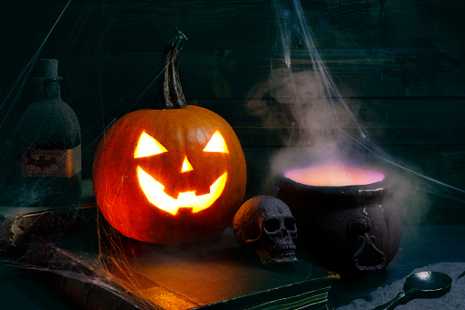 Photo courtesy of unsplash. Carved pumpkin Halloween decoration covered in spider webs and cauldron with smoke.